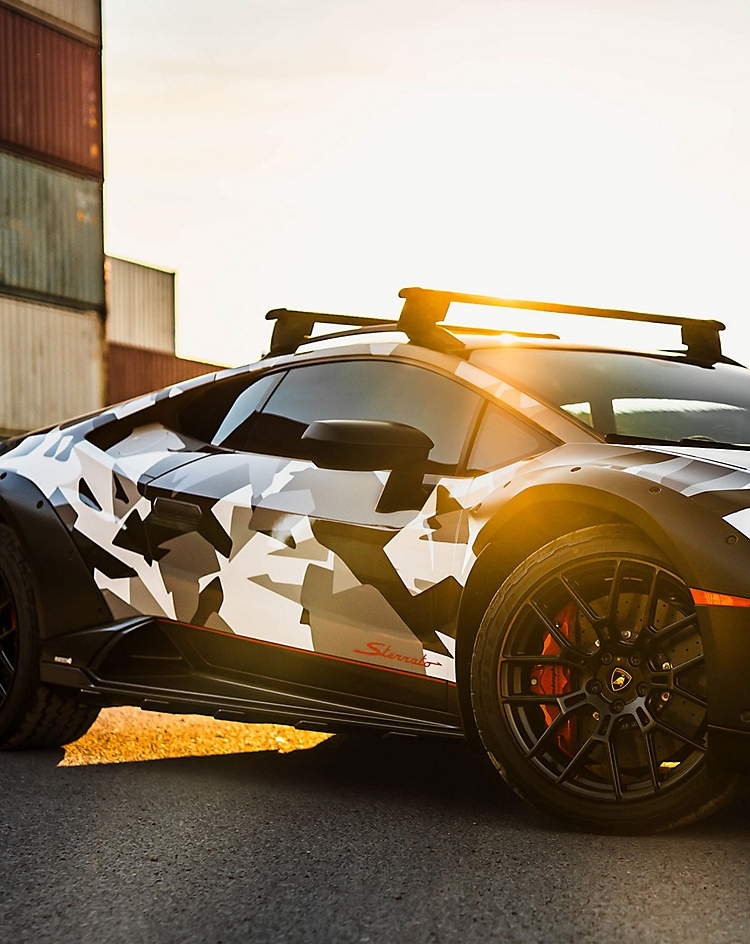 Camouflage-wrapped sports car parked near shipping containers at sunset.