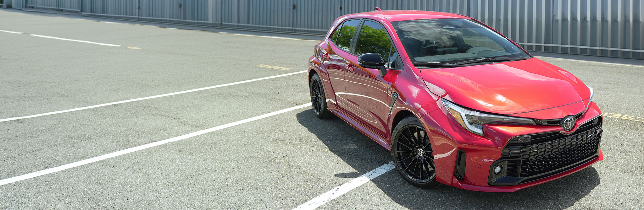 Κόκκινο hatchback σε χώρο στάθμευσης με λευκές γραμμές και μεταλλικό κτίριο στο φόντο.